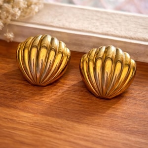 May include: Two gold-colored, shell-shaped earrings sit on a wooden surface. The earrings have a ribbed design, catching the light and creating highlights. The background includes a window and dried flowers.