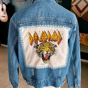 May include: A blue denim jacket featuring a large rectangular cream-colored patch on the back. The patch displays the text Def Leppard in yellow and red letters above a graphic illustration of a snarling leopard head with yellow and black fur.