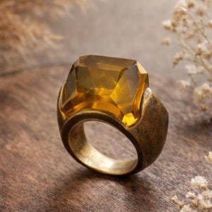 May include: A large, antique-style ring with a faceted, amber-colored gemstone. The ring's band and setting have a tarnished, bronze-like finish. The gemstone is rectangular with multiple facets, catching the light.