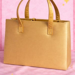 May include: A gold-colored satin handbag with two matching handles. The rectangular bag has a clasp closure and is set against a pink background with yellow star accents. The bag is a classic style.
