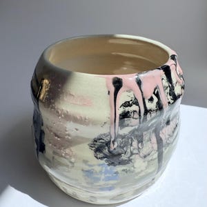Puede incluir: Un jarrón de cerámica con forma redondeada y una abertura ancha. El exterior presenta un diseño texturizado y en capas con pintura rosa y negra goteando sobre una base crema y azul pálido. El interior es de color beige claro.
