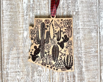 May include: Wooden ornament in the shape of Arizona, featuring a detailed etched design of desert flora and fauna. The ornament includes cacti, flowers, and a bird, with a red and black plaid ribbon for hanging. The background is a light wood.