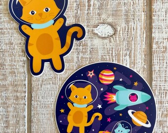 Puede incluir: Dos pegatinas con gatos naranjas con trajes espaciales. Una pegatina muestra un gato flotando en el espacio con un cohete, un OVNI, planetas y estrellas. La otra pegatina muestra un gato con un traje espacial sobre un fondo azul.