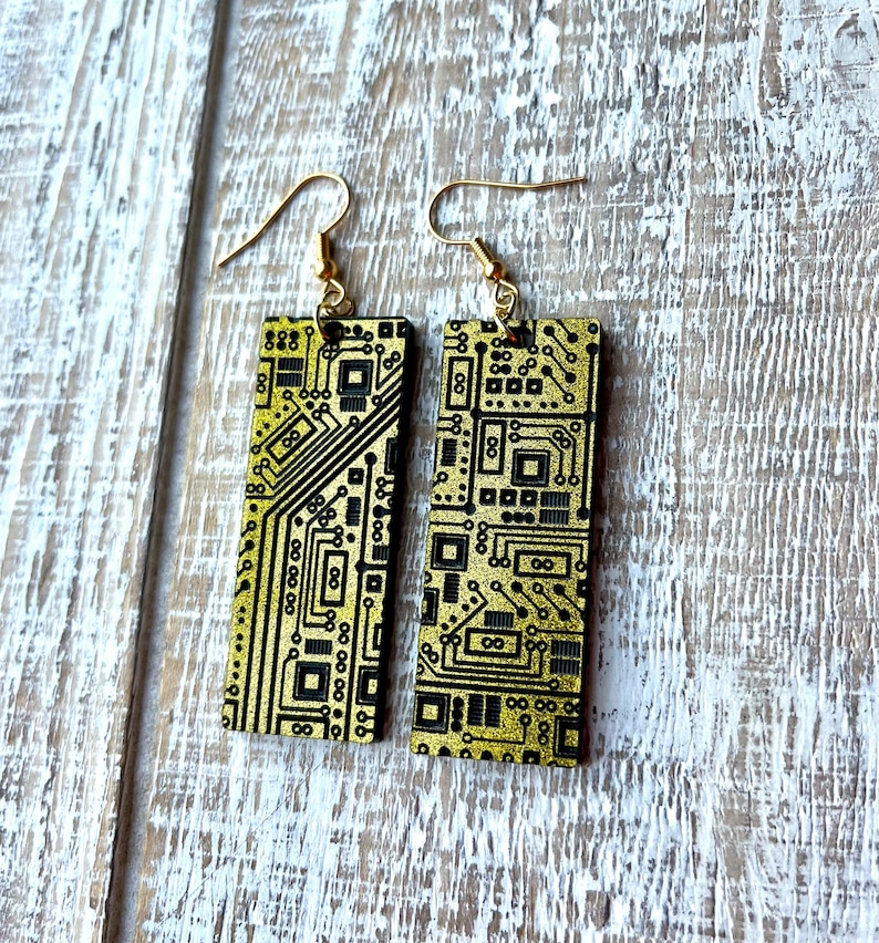 Op de afbeelding: Een paar gouden en zwarte printplaat oorbellen. De oorbellen zijn rechthoekig en hebben een gouden glitter afwerking. Het printplaat ontwerp is zwart en zichtbaar aan beide zijden van de oorbellen.