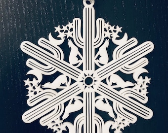 May include: White snowflake ornament with a desert theme. The design features cacti, howling coyotes, stars, and crescent moons. The ornament is suspended by a silver ribbon. The background is a dark, textured surface.