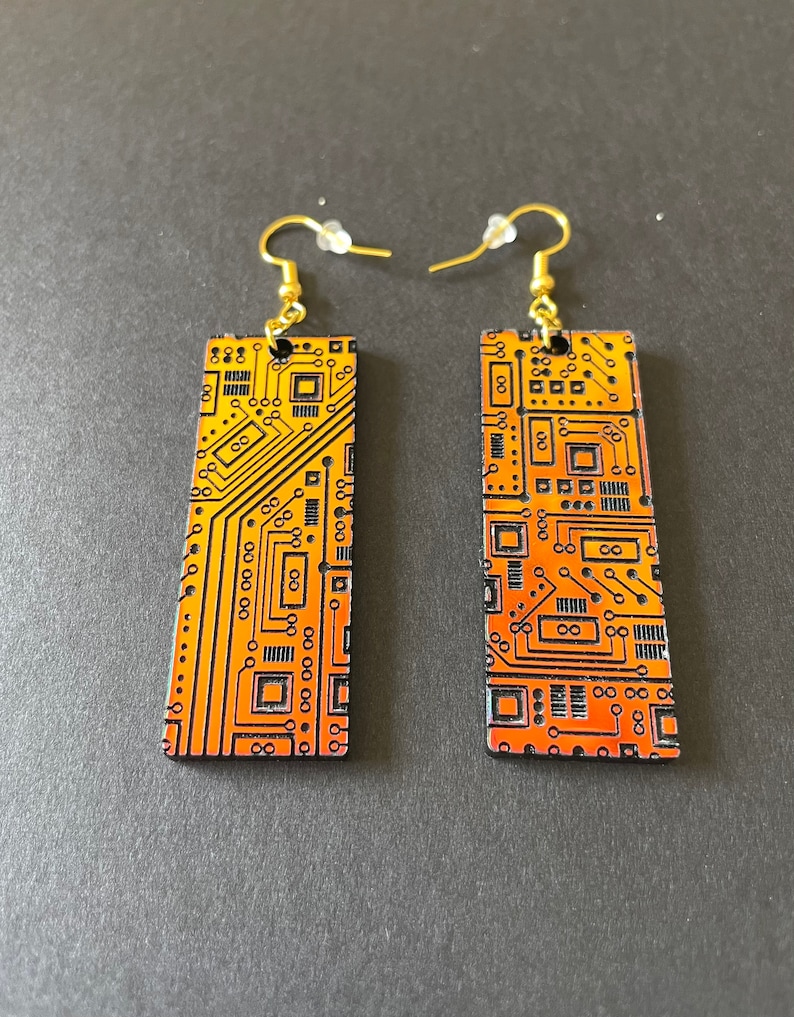 Op de afbeelding: Een paar gouden oorbellen met een rechthoekig ontwerp met een print van een printplaat in oranje en zwarte tinten.