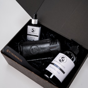 Puede incluir: Una caja de regalo negra que contiene productos para el cuidado de la barba. Incluye un cepillo para barba, aceite para barba, lavado de barba y cara y un peine con estuche de cuero. Los productos tienen etiquetas blancas con un logotipo y texto.
