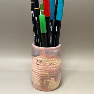 May include: A ceramic pen holder with a pink and tan color scheme, holding several colorful markers. The markers include red, green, blue, and black, with the brand name "Tombow ABT" visible on the pens. The holder has a textured surface with star and text details.