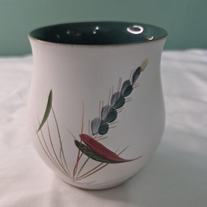 May include: A white ceramic mug with a dark green interior and a painted floral design. The design features green, red, and blue-grey leaves and stems. The mug has a rounded shape and a slightly flared rim.
