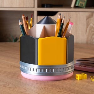 Puede incluir: Un organizador de escritorio con forma de l&aacute;piz en negro, amarillo y rosa, lleno de l&aacute;pices y pinceles. El organizador tiene una banda plateada y una base rosa. Un sacapuntas amarillo y un clip est&aacute;n sobre la superficie de madera.
