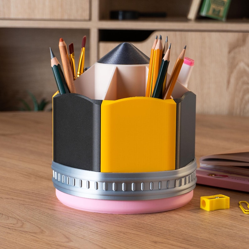 Puede incluir: Un organizador de escritorio con forma de l&aacute;piz en negro, amarillo y rosa, lleno de l&aacute;pices y pinceles. El organizador tiene una banda plateada y una base rosa. Un sacapuntas amarillo y un clip est&aacute;n sobre la superficie de madera.