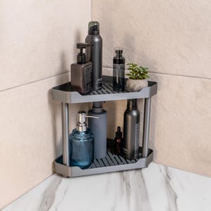 Puede incluir: Una estantería esquinera gris de dos niveles llena de varios productos de baño. La estantería contiene un dispensador de jabón de vidrio azul, botellas oscuras y una pequeña planta en maceta. La estantería está colocada en una esquina de baño con paredes de azulejos beige.