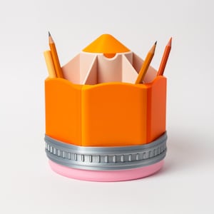 Puede incluir: Un original organizador de escritorio con forma de l&aacute;piz naranja con base rosa y ribete gris. La secci&oacute;n superior est&aacute; dividida en compartimentos, que contienen l&aacute;pices y bol&iacute;grafos. El dise&ntilde;o es una versi&oacute;n l&uacute;dica de un material escolar cl&aacute;sico.