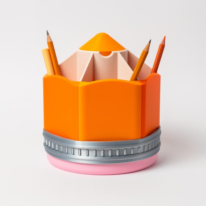 Puede incluir: Un original organizador de escritorio con forma de l&aacute;piz naranja con base rosa y ribete gris. La secci&oacute;n superior est&aacute; dividida en compartimentos, que contienen l&aacute;pices y bol&iacute;grafos. El dise&ntilde;o es una versi&oacute;n l&uacute;dica de un material escolar cl&aacute;sico.