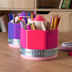Puede incluir: Dos organizadores de escritorio con forma de l&aacute;piz en colores brillantes. Uno es morado, el otro rosa. Ambos tienen una banda plateada alrededor de la base y est&aacute;n llenos de bol&iacute;grafos, l&aacute;pices y &uacute;tiles de papeler&iacute;a. Los organizadores est&aacute;n sobre un escritorio de madera.