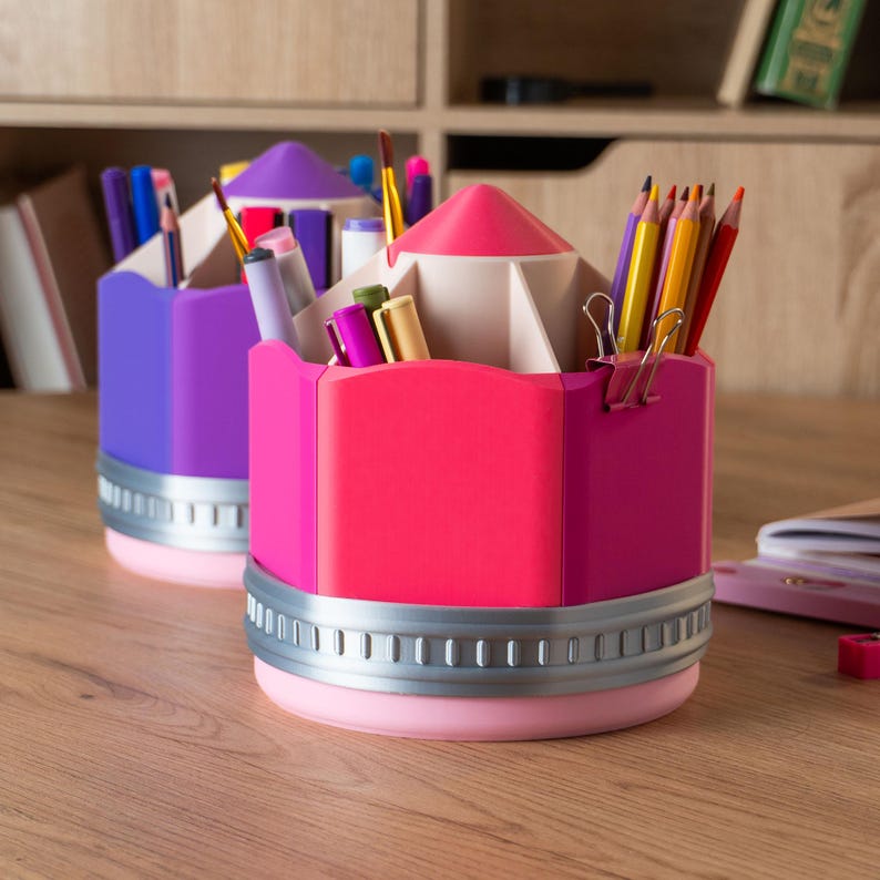 Puede incluir: Dos organizadores de escritorio con forma de l&aacute;piz en colores brillantes. Uno es morado, el otro rosa. Ambos tienen una banda plateada alrededor de la base y est&aacute;n llenos de bol&iacute;grafos, l&aacute;pices y &uacute;tiles de papeler&iacute;a. Los organizadores est&aacute;n sobre un escritorio de madera.