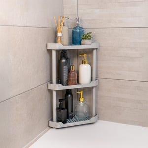 Może przedstawiać: Trzypoziomowa, szara półka narożna wypełniona produktami do łazienki. Na górnej półce znajduje się niebieski dozownik mydła, mała roślina doniczkowa i szklana butelka z trzciną. Środkowa i dolna półka prezentują różne butelki i dozowniki.