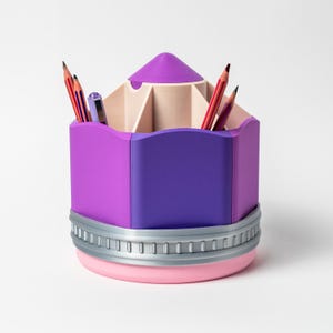 Puede incluir: Un organizador de escritorio con forma de l&aacute;piz morado y rosa con una banda plateada. El organizador contiene l&aacute;pices y bol&iacute;grafos. La parte superior es de color morado claro y la base es rosa. Dise&ntilde;ado para guardar bol&iacute;grafos y l&aacute;pices.