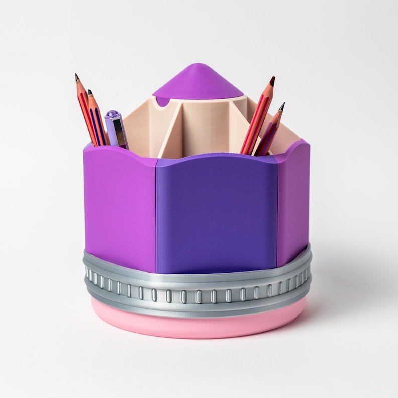 Puede incluir: Un organizador de escritorio con forma de l&aacute;piz morado y rosa con una banda plateada. El organizador contiene l&aacute;pices y bol&iacute;grafos. La parte superior es de color morado claro y la base es rosa. Dise&ntilde;ado para guardar bol&iacute;grafos y l&aacute;pices.