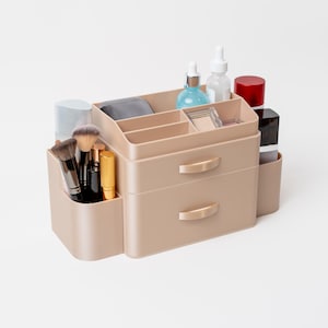 Op de afbeelding: Een beige make-up organizer met twee lades en meerdere compartimenten. Het bevat diverse beautyproducten, waaronder kwasten, lippenstiften en flesjes. De organizer heeft goudkleurige handgrepen en is ontworpen om cosmetica op te bergen en te presenteren.