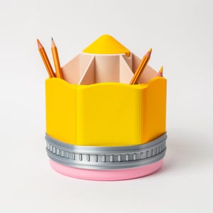 Puede incluir: Un organizador de escritorio con forma de l&aacute;piz amarillo con base rosa y ribete plateado. El organizador contiene varios l&aacute;pices naranjas. La parte superior del organizador es de color beige claro con varios compartimentos.