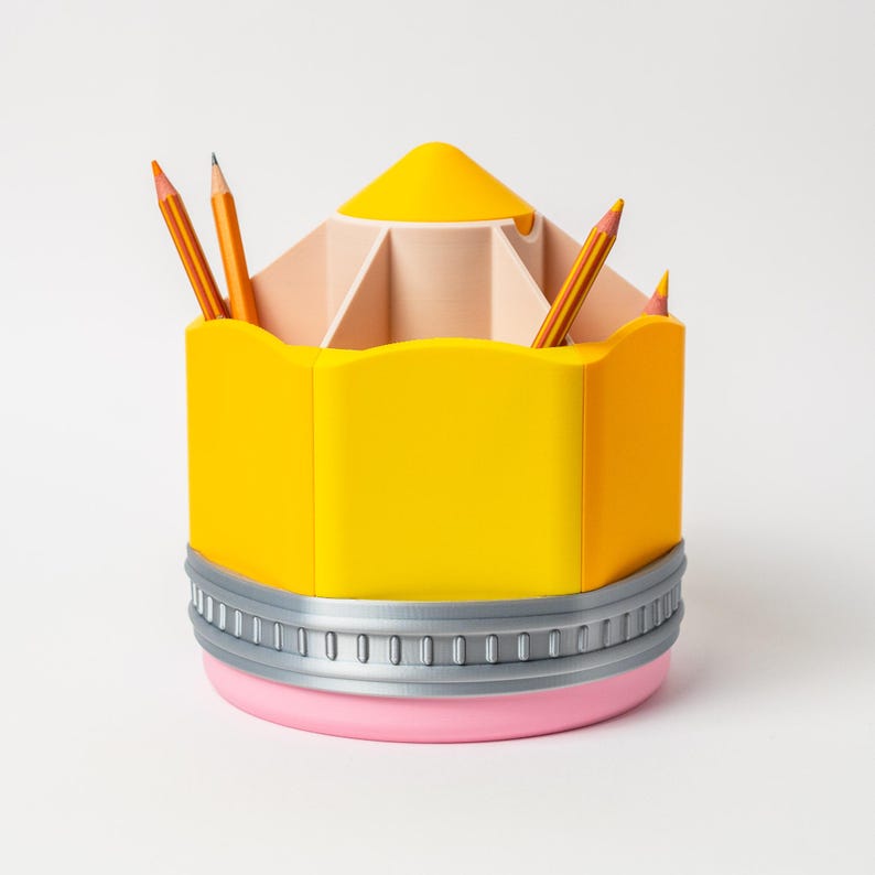 Puede incluir: Un organizador de escritorio con forma de l&aacute;piz amarillo con base rosa y ribete plateado. El organizador contiene varios l&aacute;pices naranjas. La parte superior del organizador es de color beige claro con varios compartimentos.