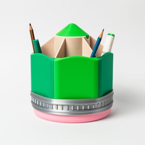 Puede incluir: Un organizador de escritorio con forma de l&aacute;piz verde con base rosa y ribete plateado. El organizador contiene l&aacute;pices y un rotulador. La parte superior es de color beige claro con separadores verdes.