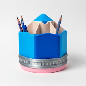 Puede incluir: Un organizador de escritorio azul con forma de l&aacute;piz, con una base rosa claro y una banda plateada. El organizador tiene m&uacute;ltiples compartimentos y contiene varios l&aacute;pices. La parte superior es azul claro. Un accesorio de escritorio divertido y funcional.