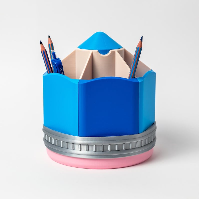 Puede incluir: Un organizador de escritorio azul con forma de l&aacute;piz, con una base rosa claro y una banda plateada. El organizador tiene m&uacute;ltiples compartimentos y contiene varios l&aacute;pices. La parte superior es azul claro. Un accesorio de escritorio divertido y funcional.