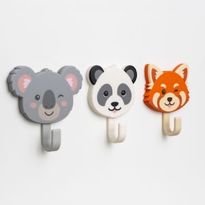 Pode incluir: Três ganchos de parede em forma de animais: um coala cinzento, um panda e um panda vermelho. Cada gancho tem um rosto sorridente e um gancho curvo por baixo. O coala pisca. O panda tem orelhas e manchas pretas. O panda vermelho pisca.