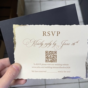 Può includere: Un biglietto di RSVP con un bordo dorato e un bordo strappato. La carta include il testo "RSVP", "Kindly reply by June 26th", un codice QR e un indirizzo web. Una busta blu navy è dietro la carta.