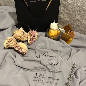 May include: A wedding invitation with the text "Romeo & Juliet" and event details. The invitation is accompanied by decorative candles, dried roses, and a black card with gold lettering. The invitation is on a gray fabric.