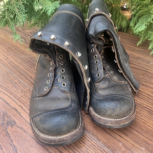 Pode incluir: Um par de botas de couro preto com biqueira arredondada, sola de borracha grossa e cano alto com ilhós de metal prateado e tachas ao longo do colarinho superior. As botas apresentam sinais de desgaste com superfícies arranhadas e couro gasto.