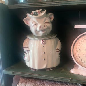 May include: A ceramic cookie jar shaped like a pig wearing a hat and a collared coat. The pig has closed eyes, long eyelashes, and a smiling expression. The jar is primarily white with green, brown, and red painted details on the hat, collar, and coat buttons.