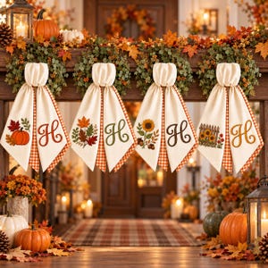 May include: Four cream-coloured fabric towels with orange gingham trim, each featuring an embroidered autumn motif and a large script letter H. The designs include a pumpkin, fall leaves, a sunflower, and a sunflower garden with a fence.