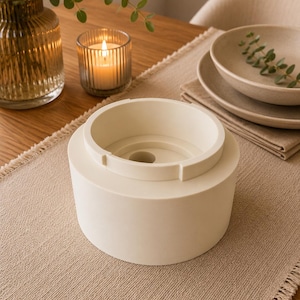 May include: A cylindrical off-white object with a stepped top rim and a central circular opening. The item features a smooth, matte finish and a tiered structure, with a smaller upper section rising from a wider, solid base, creating a distinct geometric profile.