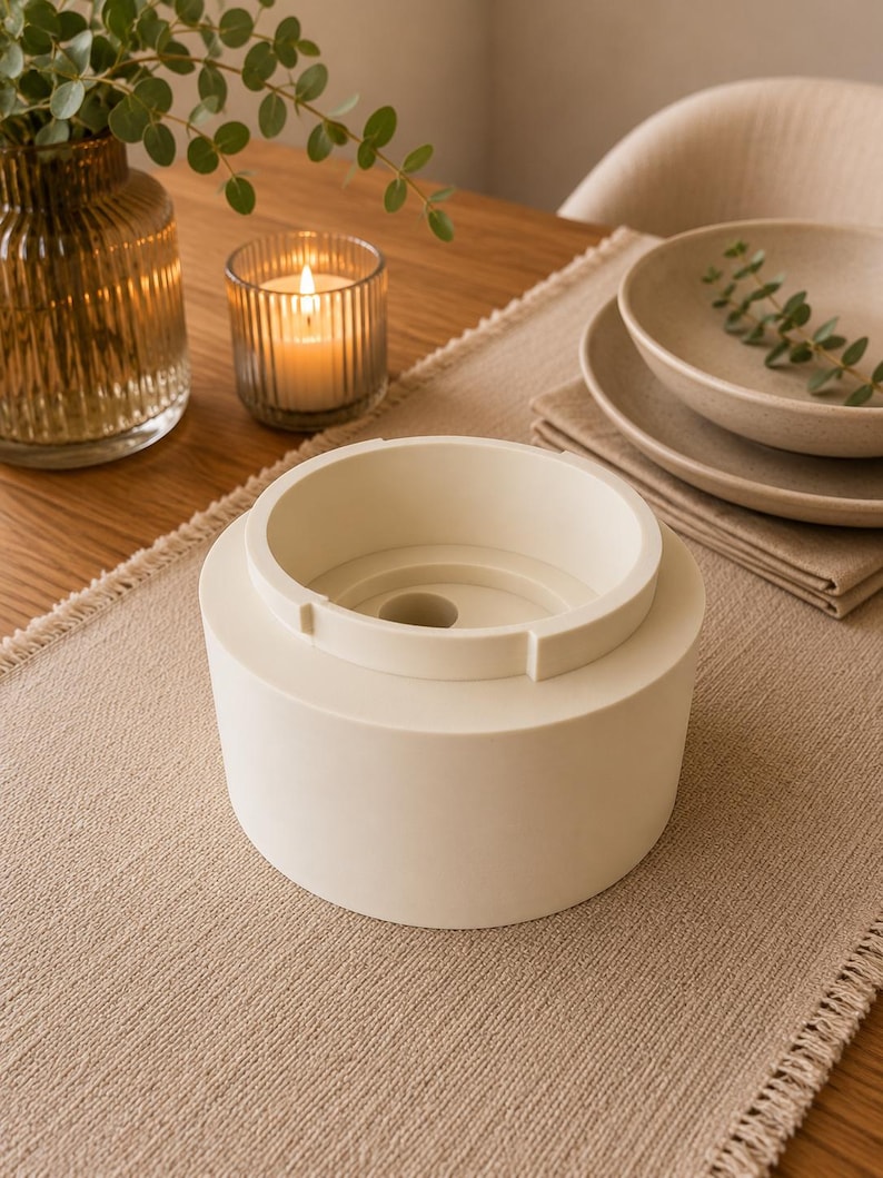May include: A cylindrical off-white object with a stepped top rim and a central circular opening. The item features a smooth, matte finish and a tiered structure, with a smaller upper section rising from a wider, solid base, creating a distinct geometric profile.