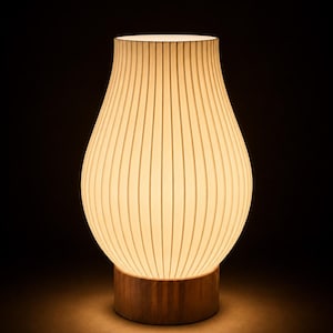 May include: A table lamp with a pear-shaped, off-white, vertically ribbed shade that emits a warm, soft glow. The shade is supported by a cylindrical, medium-brown wooden base with a visible grain, creating a contrast between the light and dark elements.
