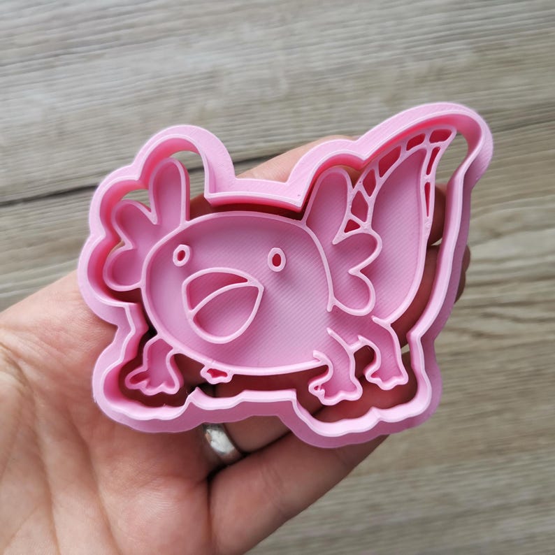 K&ouml;nnte beinhalten: Ein rosa Ausstecher in Form eines Axolotls. Der Ausstecher zeigt einen l&auml;chelnden Axolotl mit detaillierten Kiemen und einem wellenf&ouml;rmigen Schwanz. Der Ausstecher wird in einer Hand vor einem Holzgrund gehalten.
