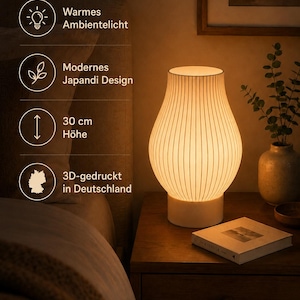 May include: A white table lamp with a ribbed, vase-shaped shade emitting a warm glow. The lamp stands on a cylindrical base. Text on the image indicates: Warmes Ambientelicht, Modernes Japandi Design, 30 cm H&ouml;he, 3D-gedruckt in Deutschland.