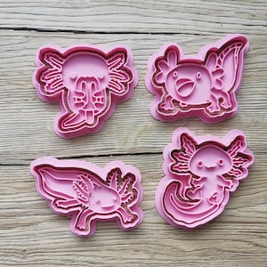 K&ouml;nnte beinhalten: Vier rosa Ausstecher in Axolotl-Form. Jeder Ausstecher zeigt ein anderes Axolotl-Design mit Details wie Kiemen und l&auml;chelnden Gesichtern. Die Ausstecher sind aus Kunststoff und auf einer Holzoberfl&auml;che angeordnet.