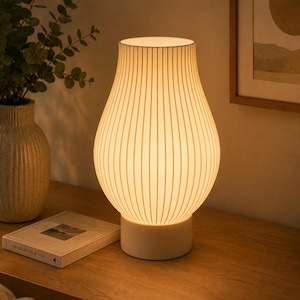 May include: A table lamp with a white, ribbed, pear-shaped shade that emits a warm, soft glow. The lamp features a solid, cylindrical white base and a vertical line pattern across the surface of the shade, creating a textured appearance.