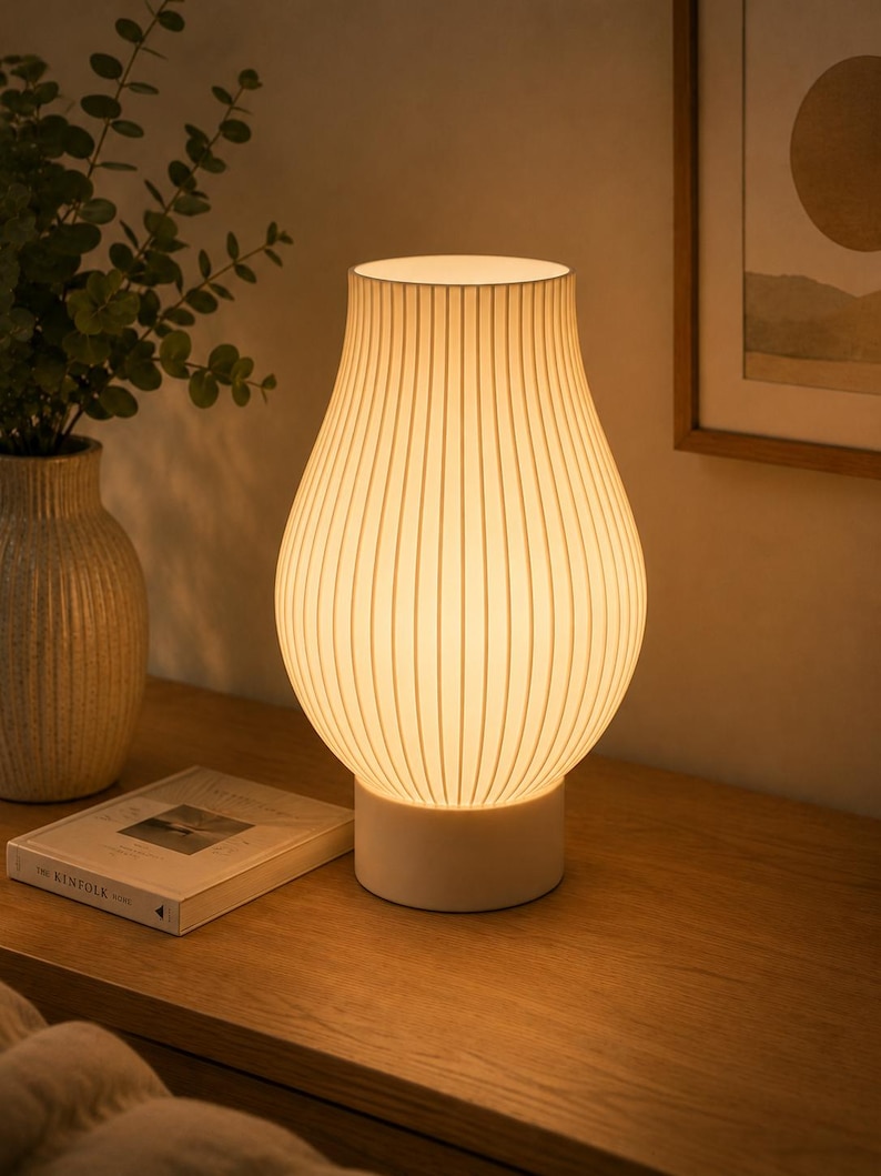 May include: A table lamp with a white, ribbed, pear-shaped shade that emits a warm, soft glow. The lamp features a solid, cylindrical white base and a vertical line pattern across the surface of the shade, creating a textured appearance.