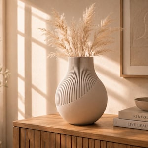 Könnte beinhalten: Eine matte, cremeweiße Vase mit rundem Boden und vertikaler Rippenstruktur in der oberen Hälfte. Die Vase ist mit getrocknetem beigem Pampasgras gefüllt. Daneben liegen zwei gestapelte Bücher mit der Aufschrift KINFOLK und LIVE BEAUTIFULLY sowie eine kleine Schale.