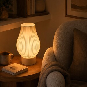 May include: A table lamp with a curved, white, ribbed glass shade that emits a warm, soft glow. The lamp features a solid, cylindrical white base and a slender, tapered neck, providing a diffused light source for a cozy, illuminated atmosphere.