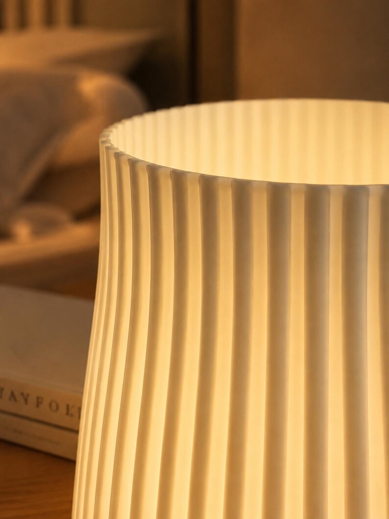 May include: A cylindrical lamp shade with a vertical ribbed texture in an off-white color. The shade is illuminated from within, casting a warm, soft glow through the translucent material, highlighting the repeating pattern of the vertical ridges.