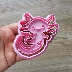 K&ouml;nnte beinhalten: Ein rosa Ausstecher in Form eines Axolotl. Der Ausstecher aus Kunststoff zeigt ein detailliertes Design des Wassersalamanders. Der Axolotl hat ein l&auml;chelndes Gesicht, krause Kiemen und einen langen, flie&szlig;enden Schwanz. Zum Kekse backen.