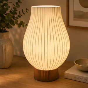 May include: A table lamp featuring a white, vertically ribbed, pear-shaped shade that emits a warm glow. The lamp is supported by a cylindrical base with a metallic finish, providing a stable foundation for the textured, translucent shade.