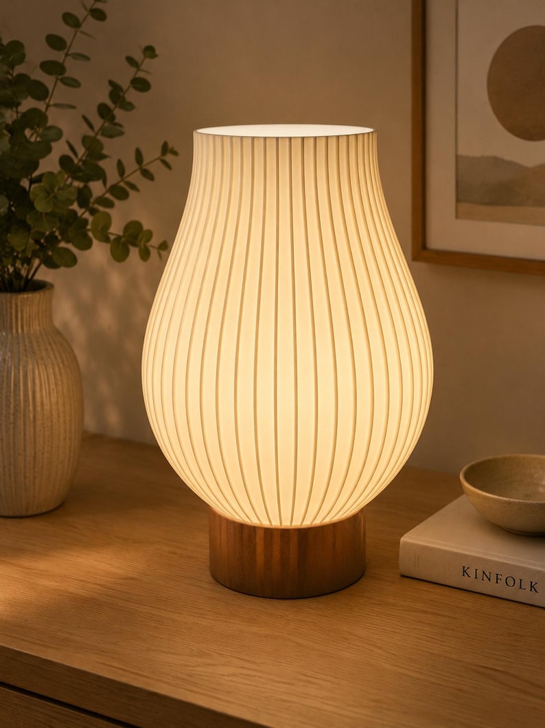 May include: A table lamp featuring a white, vertically ribbed, pear-shaped shade that emits a warm glow. The lamp is supported by a cylindrical base with a metallic finish, providing a stable foundation for the textured, translucent shade.