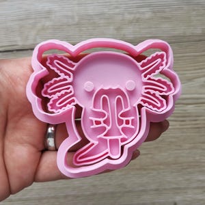 K&ouml;nnte beinhalten: Ein rosa Kunststoff-Ausstecher in Form eines Axolotls. Der Ausstecher hat die Form eines Axolotls mit detaillierten Kiemen, einem l&auml;chelnden Gesicht und einem abgerundeten Kopf. Der Ausstecher wird in einer Hand gehalten.